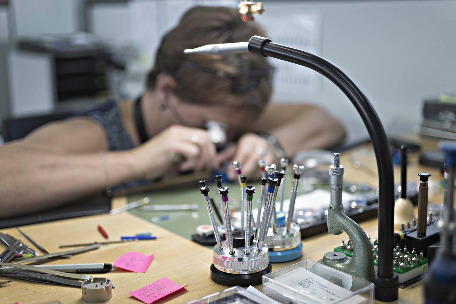 An employee uses a loupe to inspect the mechanism of a wristwatch. The tools in the foreground are what she uses to pick up or to manipulate the almost-microscopic components.